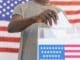 A person casting their vote at a polling station with a US flag in the background.