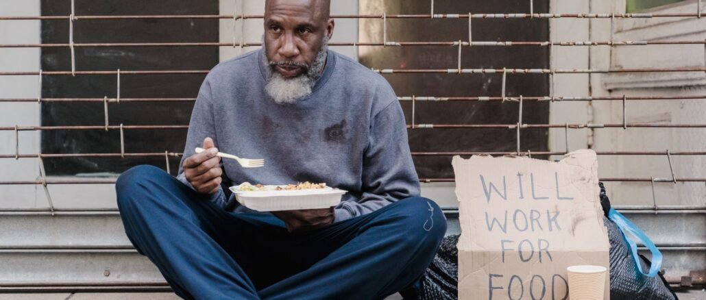 Elderly homeless man sitting on street with a sign and meal, symbolizing poverty.
