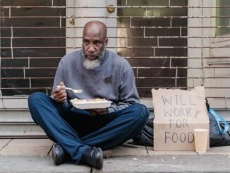 Elderly homeless man sitting on street with a sign and meal, symbolizing poverty.