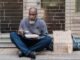 Elderly homeless man sitting on street with a sign and meal, symbolizing poverty.