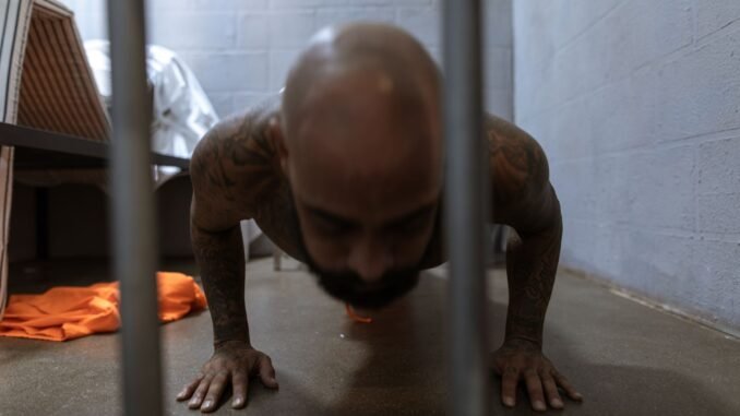 Man in prison uniform doing push-ups inside a cell with concrete walls.